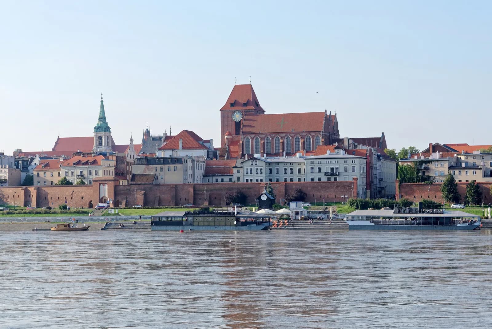 A wide view of the city of Torun, Poland, showcasing its historic architecture including a prominent church with a large red roof and clock tower, and a green-spired building, all situated alongside a river with boats in the foreground under a clear sky.