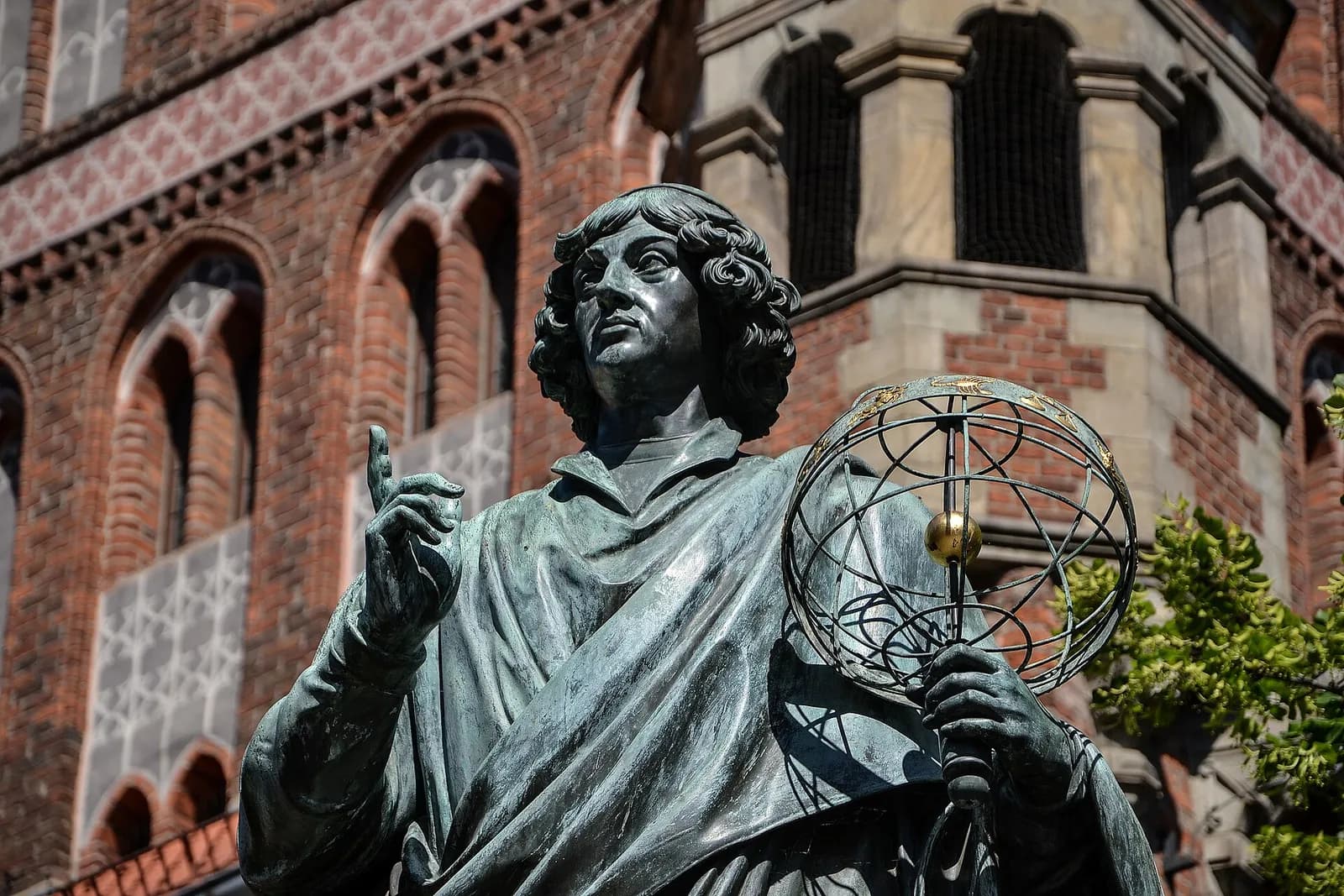 A close-up shot of a bronze statue of a man with curly hair, holding a sphere, set against an out-of-focus brick building with arched windows.
