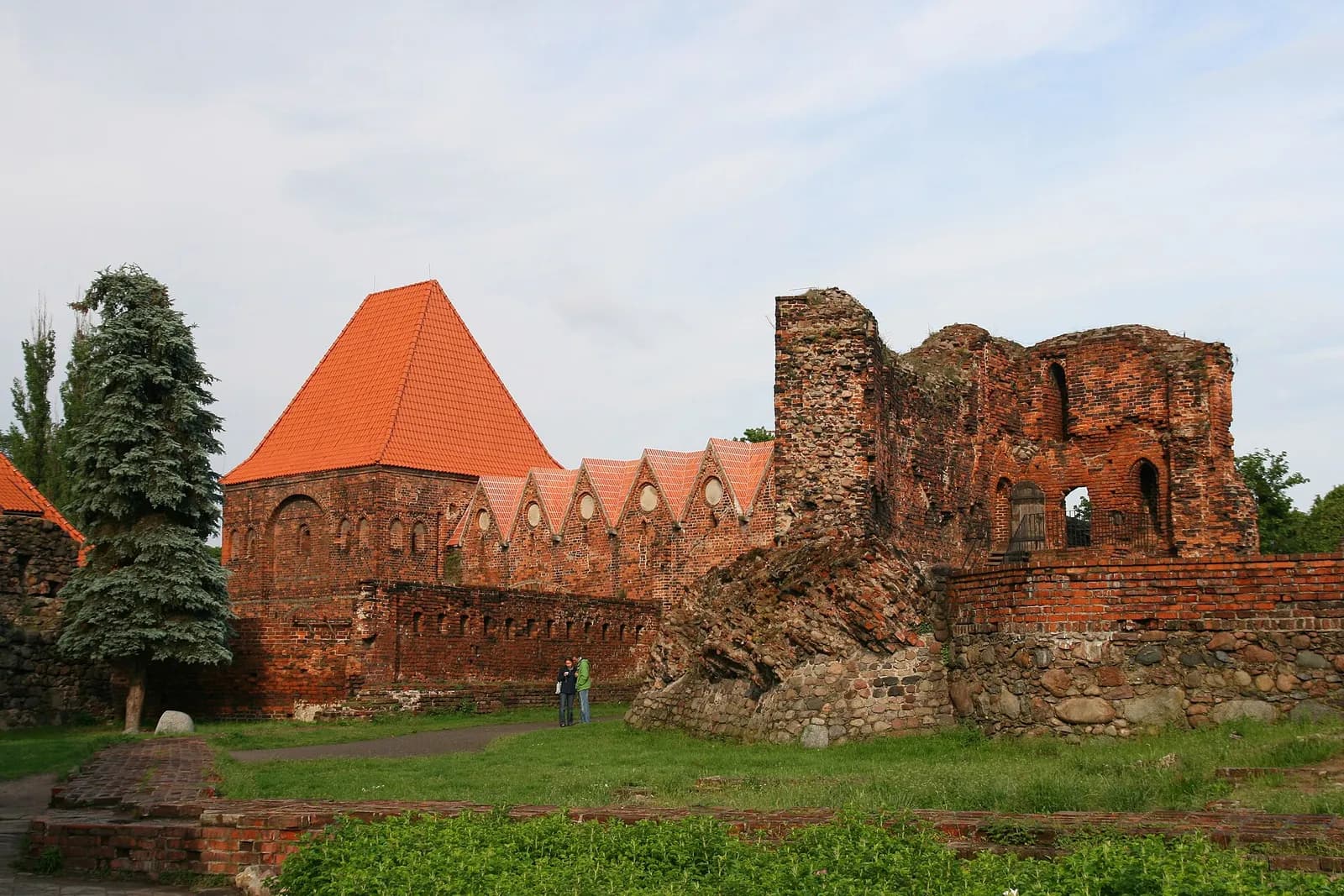 A large red brick structure featuring a prominent red tile roof on the left and a more ruined section on the right, surrounded by green grass and a tall pine tree, under an overcast sky.
